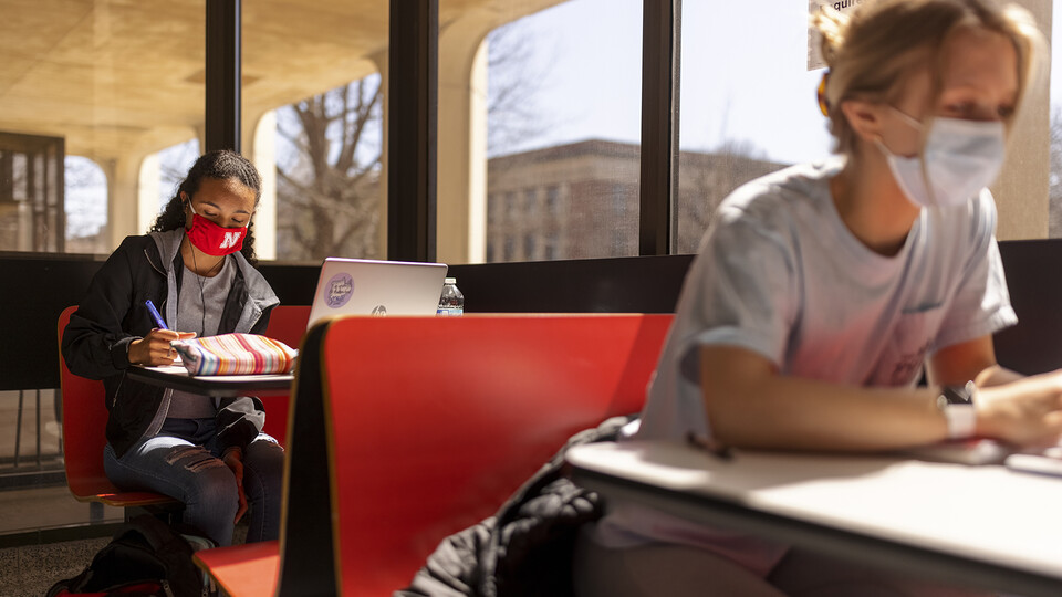 Gabriella Silva (red mask) and Zoe Keese (white mask) study at the tables in the sun lit lobby inside Hamilton Hall during the spring 2021 semester.