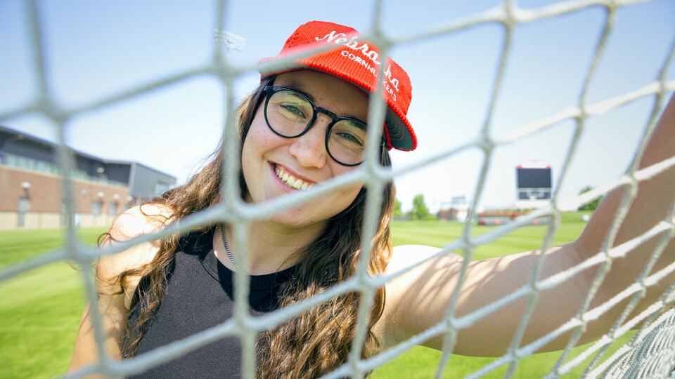 Cece Villa, Husker soccer goalkeeper, smiles for a photo through the net of the goal.