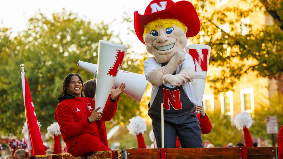 Herbie Husker and Spirit Squad member Amani Mfinanga cheer from a tractor-pulled wagon in the 2023 homecoming parade. [Craig Chandler | University Communication and Marketing]