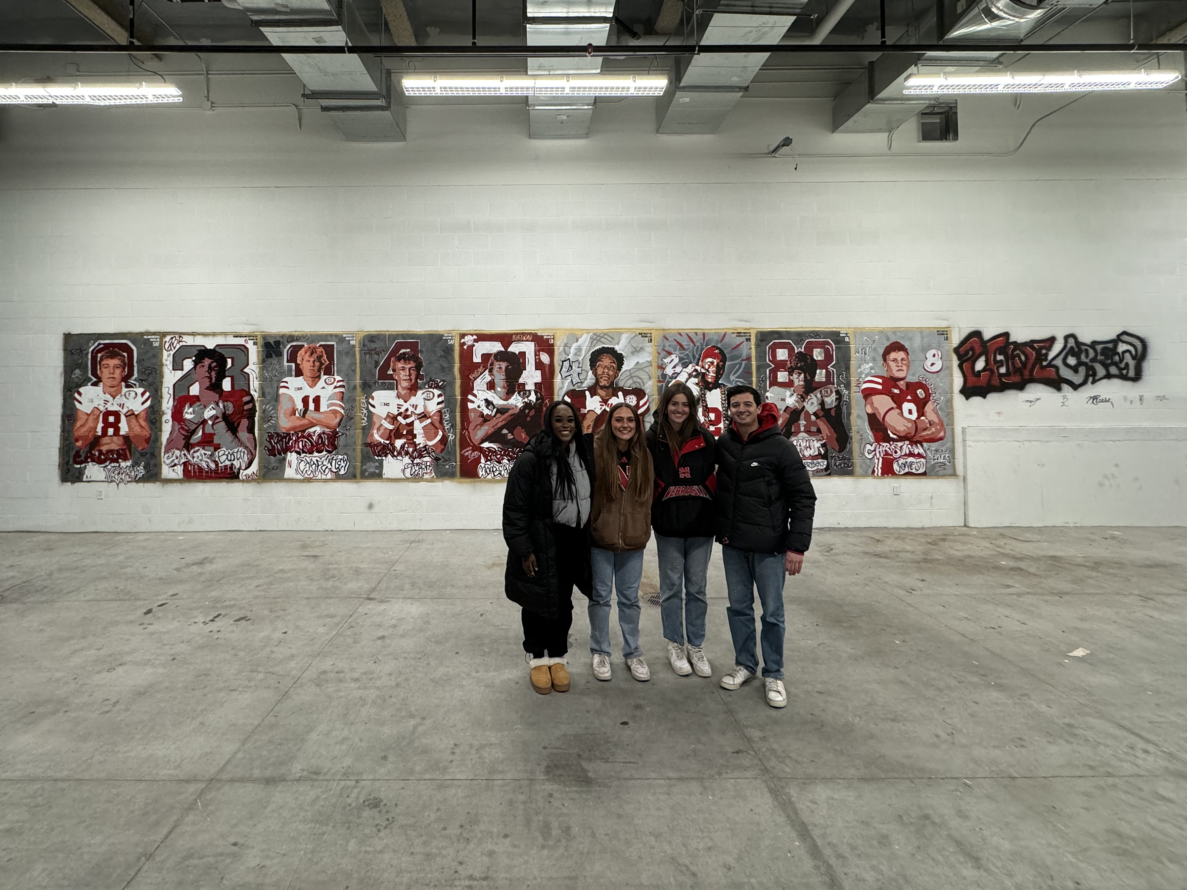 Left to right:  Michelle Reese, Maddie Vanderbur, Lea Bushey and Micah Fullinfaw stand in front of the mural they created to spotlight the players signed on National Signing Day by the Nebraska Football team. Courtesy photo.