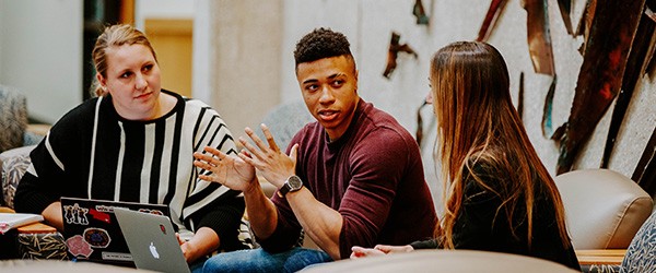 Three people sitting at a table in front of computer talking