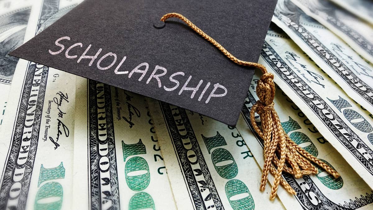 A black graduation cap with a gold tassel sits on a pile of U.S. $100 bills. The word &lsquo;Scholarship&rsquo; is written in white script across the top of the cap.