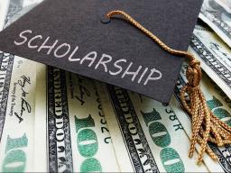 A black graduation cap with a gold tassel sits on a pile of U.S. $100 bills. The word &lsquo;Scholarship&rsquo; is written in white script across the top of the cap.