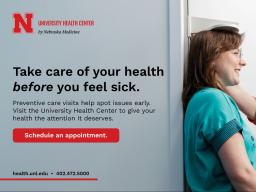 A health care staff member wearing a red Nebraska Medicine T-shirt measures a patient&rsquo;s height against a wall-mounted stadiometer, with both individuals smiling.