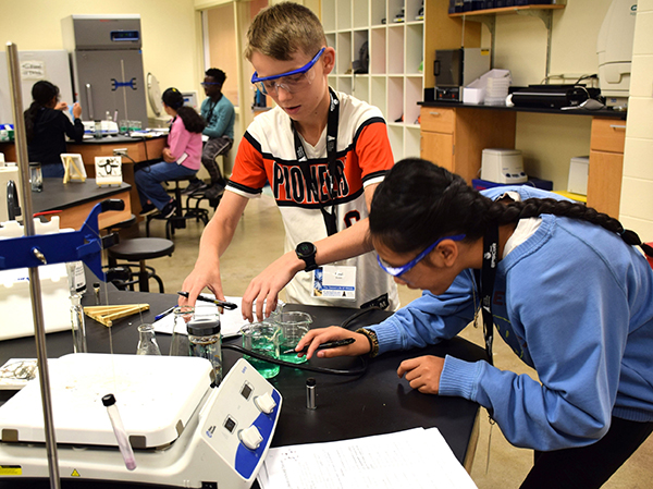 In a classroom laboratory during a Young Nebraska Scientists camp, a boy and a girl work with flasks of colored liquids while another table of students gathers in the background.
