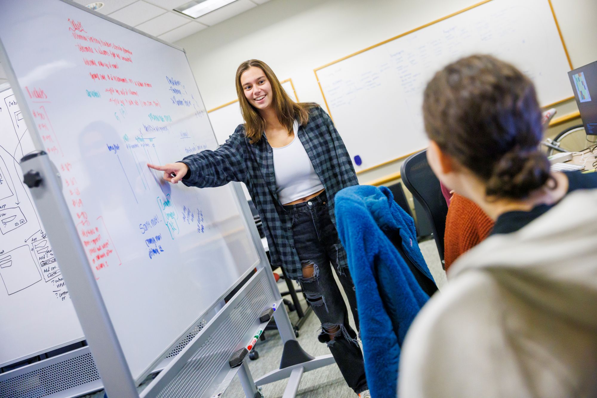 A smiling student teaches her peer while pointing to a white board.