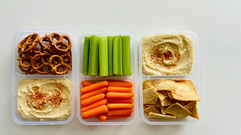 Pretzels, celery, carrots, pita chips, and hummus arranged in clear food containers.